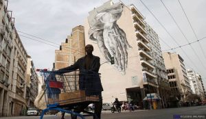 Un immigrato che vive in Grecia, spinge un carrello della spesa pieno di spazzatura, davanti a un murale dipinto su un edificio. John Kolesidis